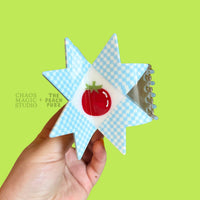 Hand holding a star-shaped object with a red tomato design against a green background, featuring 'Chaos Magic + Peach Fuzz' text.