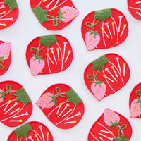 Embroidered red tomato-shaped patches with colorful pins and a pink strawberries, against a white background