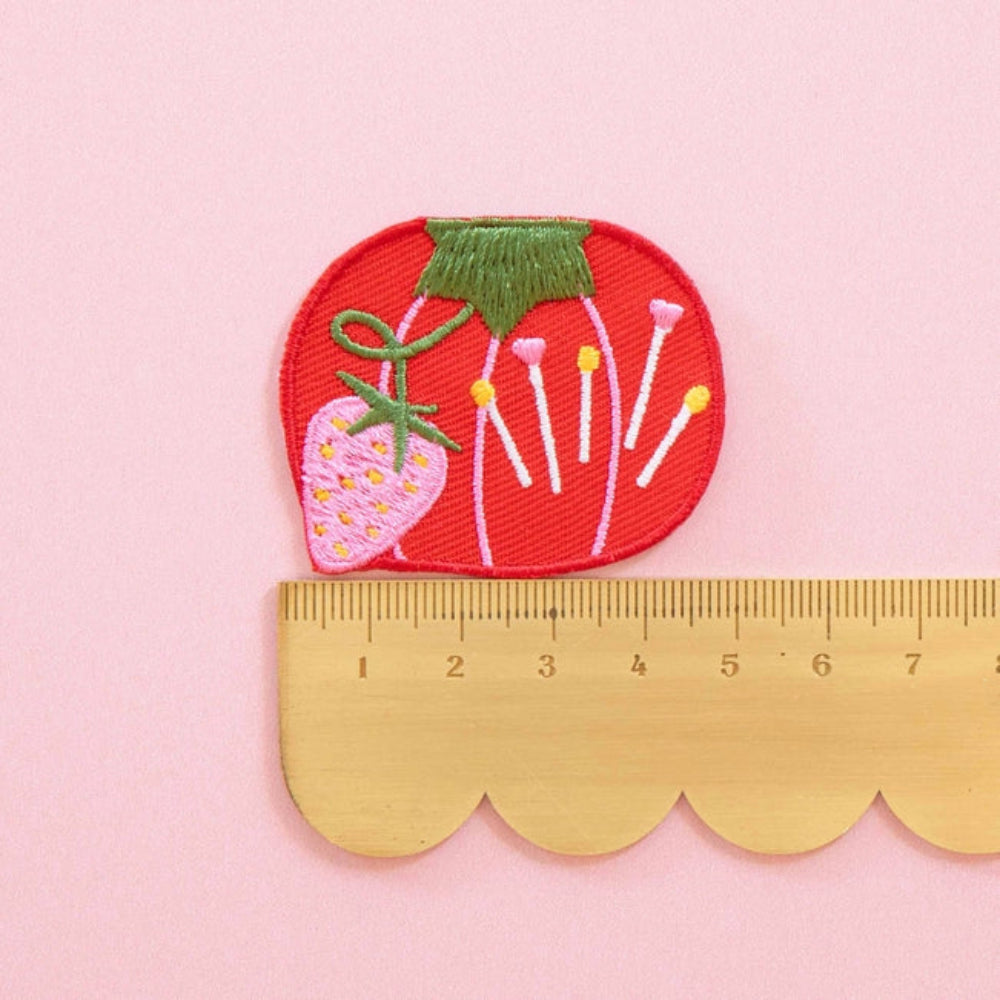 Embroidered red tomato-shaped patch with colorful pins and a pink strawberry, placed on a scalloped edge ruler against a pink background