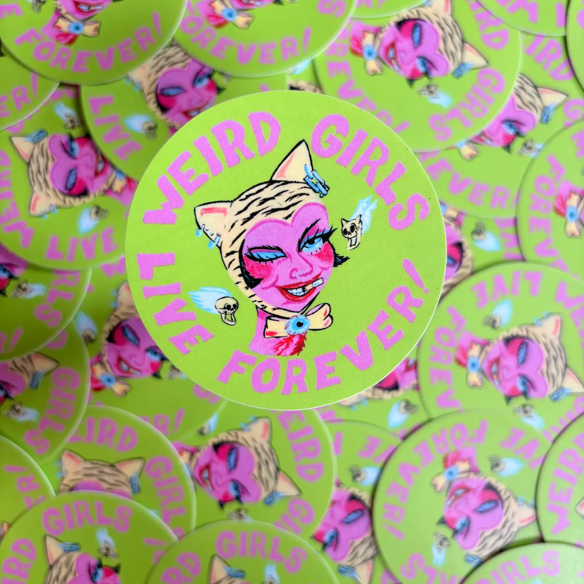 Pile of circular green stickers with the text "Weird girls live forever!" and an illustration of a girl wearing cat ears.