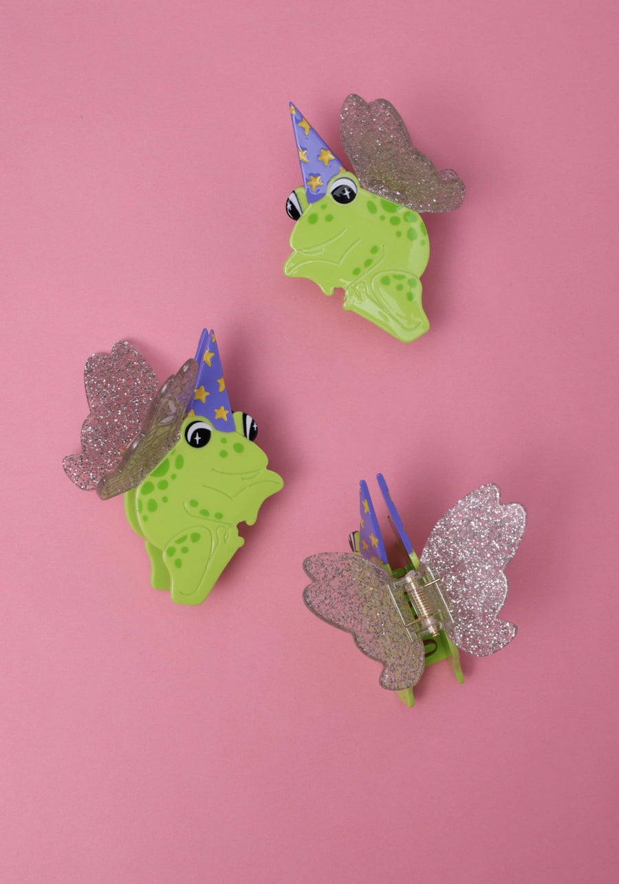 Three decorative frog clips with glittery wings and wizard hats on a pink background