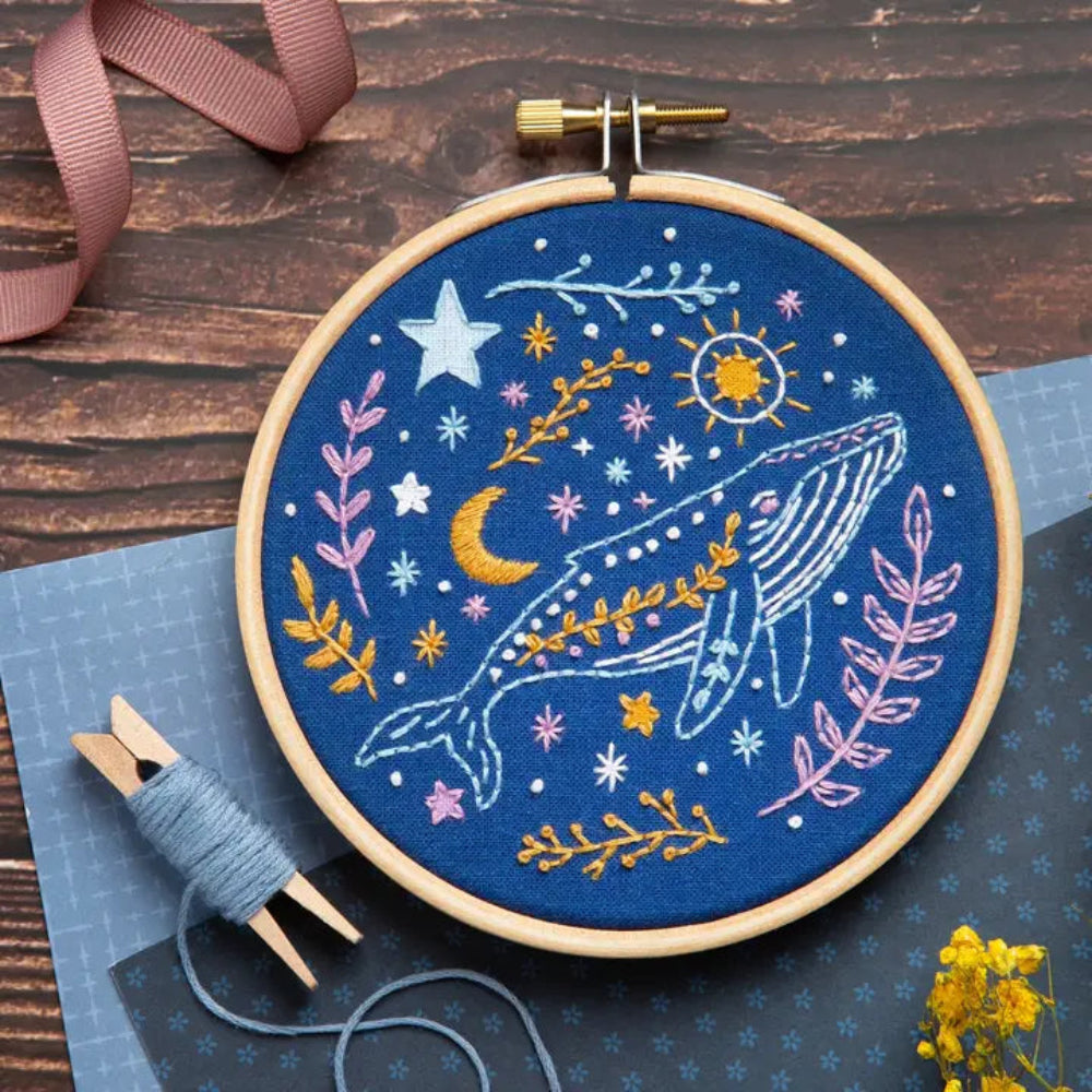 Embroidery hoop with whale design on blue fabric, surrounded by thread and ribbon on a wooden surface.