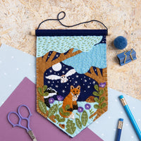 Felt pennant featuring trees, an owl, a fox, flowers, and a night sky on a textured surface