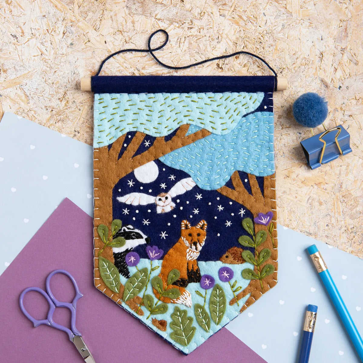 Felt pennant featuring trees, an owl, a fox, flowers, and a night sky on a textured surface