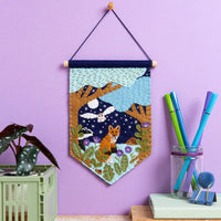 Felt pennant featuring trees, an owl, a fox, flowers, and a night sky hanging on a purple background.
