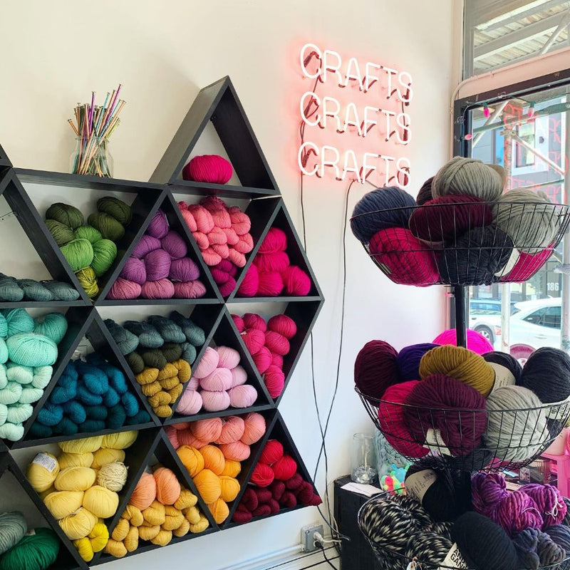 Colorful yarn on shelves with a neon sign reading 'Crafts' in a craft store.